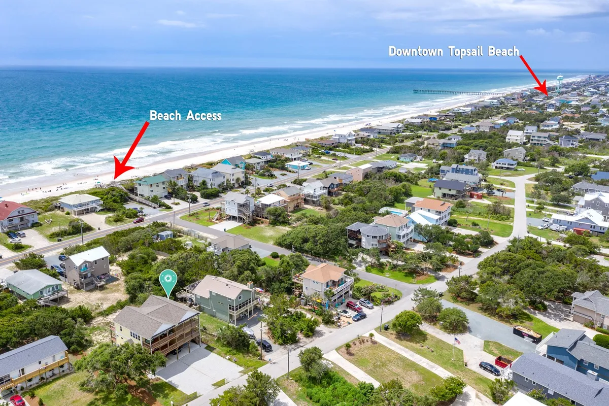 This image offers a sweeping view of the ocean, direct beach access, neighboring residential homes, and elements of downtown Topsail Beach in the distance. The scene is active, suggesting people walking, biking, and exploring local shops and attractions.