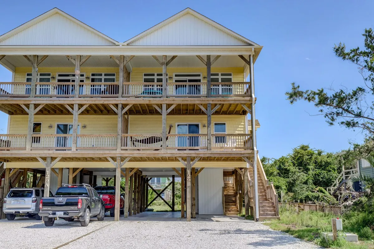 The image depicts the home's exterior, showcasing a three-story wooden house with balconies and several cars parked underneath. The ample parking space and the structure's proximity to outdoor areas are clearly visible, emphasizing easy outdoor access.