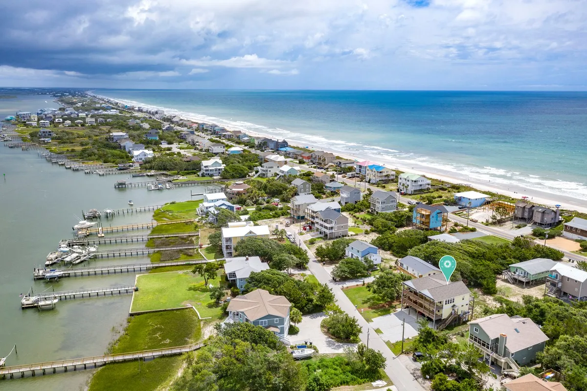 A sweeping view of the ocean with beachfront properties and a marina in sight. The image showcases sandy shores, calm blue waters, and vibrant green spaces, capturing the heart of Topsail's beachside environment. People are seen enjoying fishing, boating, and lively beach activities, highlighting the area's appeal for outdoor enjoyment and group fun.