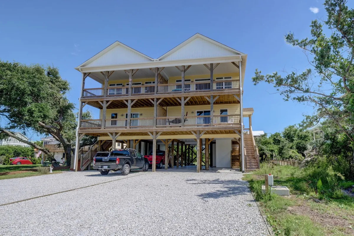 A welcoming view of a three-story house surrounded by trees, with a front porch and cars parked in the driveway. The spacious exterior hints at generous accommodations suitable for larger groups or families. The overall atmosphere is inviting, suggesting comfort and space for gathering.