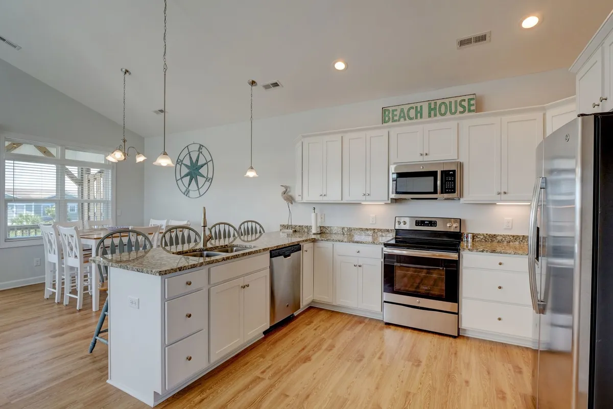 This image centers on a spacious kitchen island with granite countertops, a visible 'beach house' sign adding a touch of coastal charm, and a dining area seamlessly connected in the background. The setup hints at a kitchen designed for both serious cooking and social gatherings, with ample space and an inviting atmosphere.