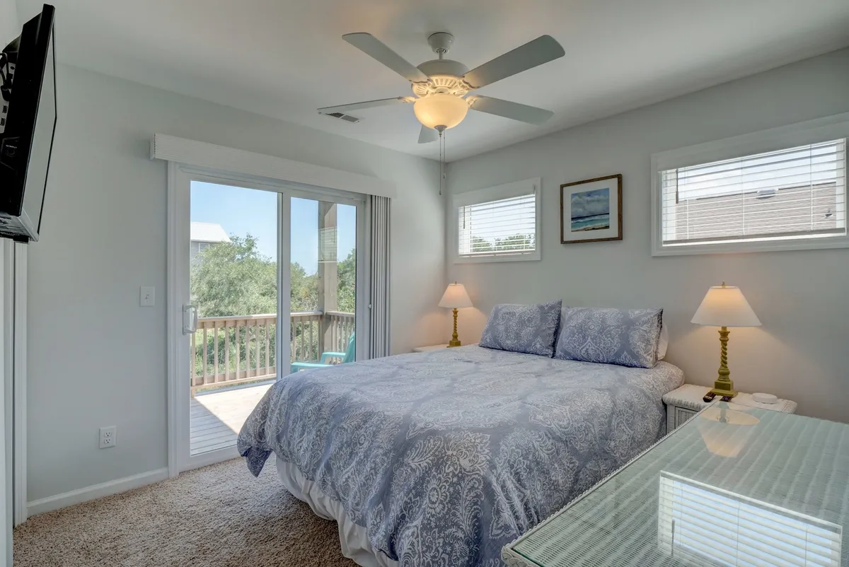 A sunlit bedroom with a bed adorned in blue patterned bedding, a glass-topped table, and a ceiling fan. The room features a balcony door, letting in abundant natural light and offering access to the outdoors.