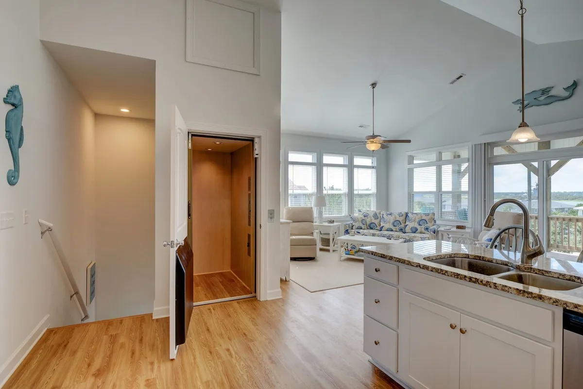 A bright, open kitchen with an island in the foreground, adjacent to a living area with large windows that let in natural light. The space feels airy and integrated, ideal for meal prep while staying connected with family and friends nearby.