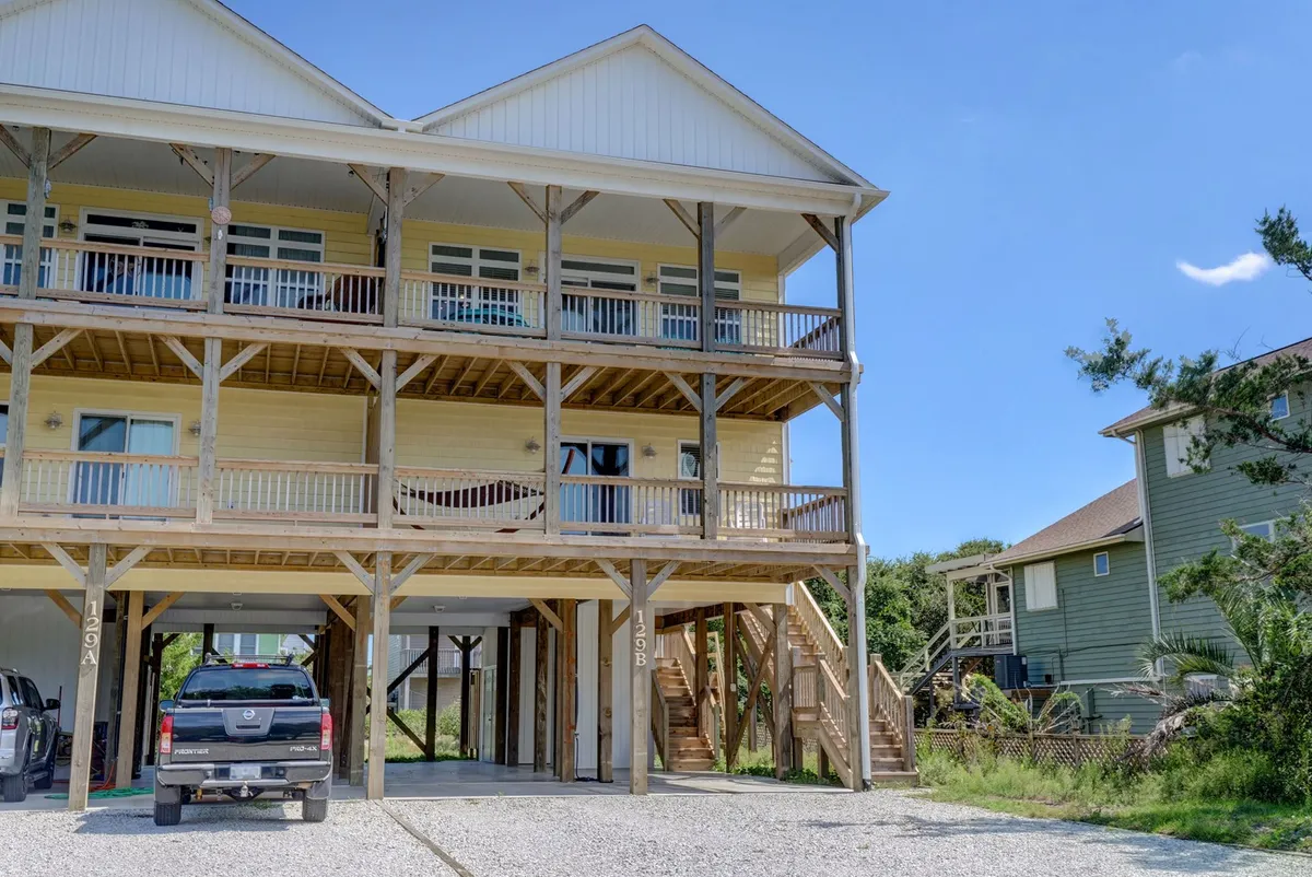 A three-story wooden beach house with multiple balconies and a prominent staircase leading upwards. The inviting exterior includes railings and open spaces ideal for gathering, relaxing, and appreciating sunrise views or evening get-togethers under the stars.