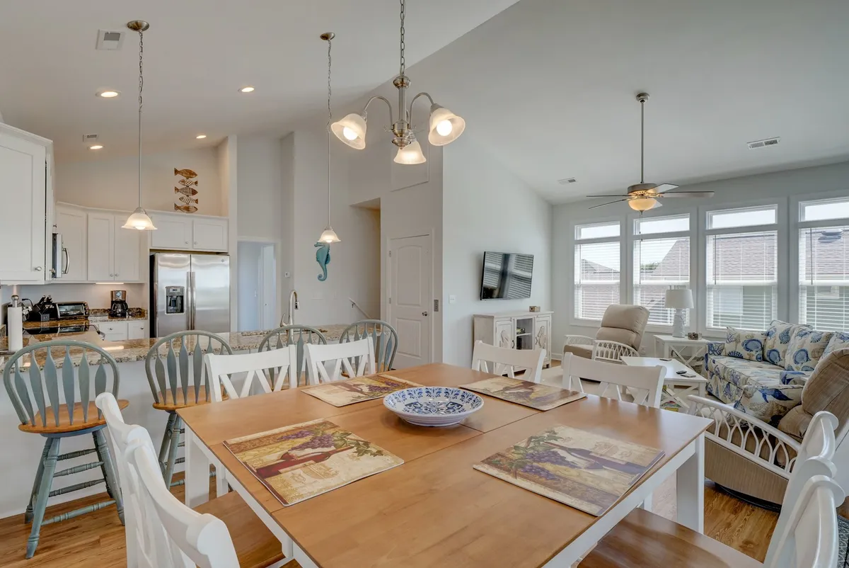 This image showcases an open-concept living space featuring a spacious dining table, a kitchen island, and a comfortable living area with a couch. Large windows flood the area with natural light, and the arrangement encourages both socializing and relaxation. The space clearly accommodates group gatherings and shared meals, as well as moments of connection and unwind after a day at the beach.
