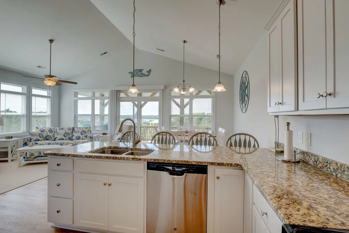 The image highlights a kitchen island with granite countertop, a dining area bathed in natural light from large windows, and a comfortable sofa. The setup is ideal for groups to gather for meals, coffee, or conversation, underscoring a modern, communal vibe.