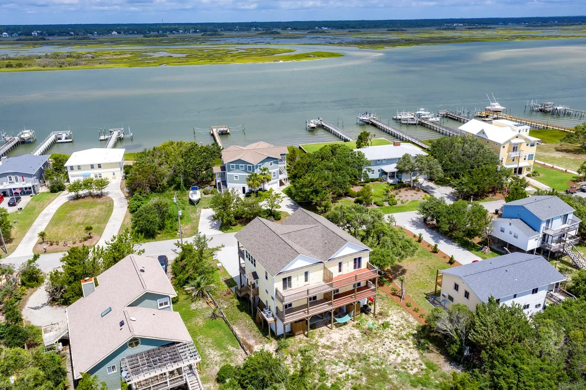 The image showcases a peaceful waterfront scene with elegant houses lining the marshland, private docks stretching into the calm waterway, and a golden sunset casting warm light over the landscape. The setting invites relaxing activities like sipping coffee on the deck, enjoying sunset views, and exploring the natural beauty by boat or on foot.
