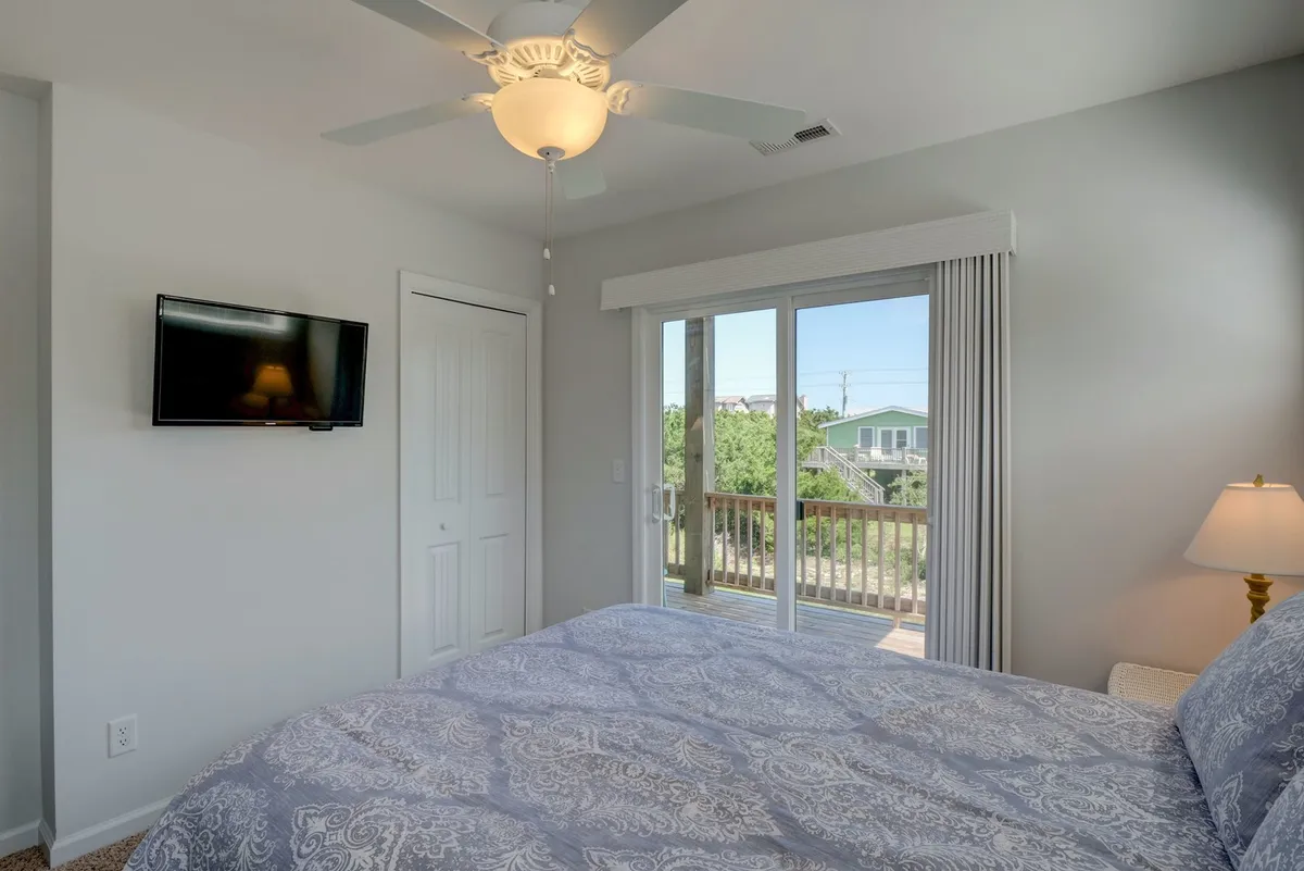 A serene bedroom featuring a bed with patterned bedding, a sliding glass door, ceiling fan, TV on the wall, and a cozy nightstand with a lamp. The space is bathed in natural light, creating a sense of calm and relaxation.