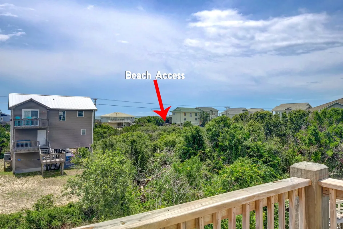 A bright, inviting exterior shot showing a beach access sign framed by leafy green foliage and neighboring houses. The pathway beckons guests to easily journey between the home and the sandy shore.